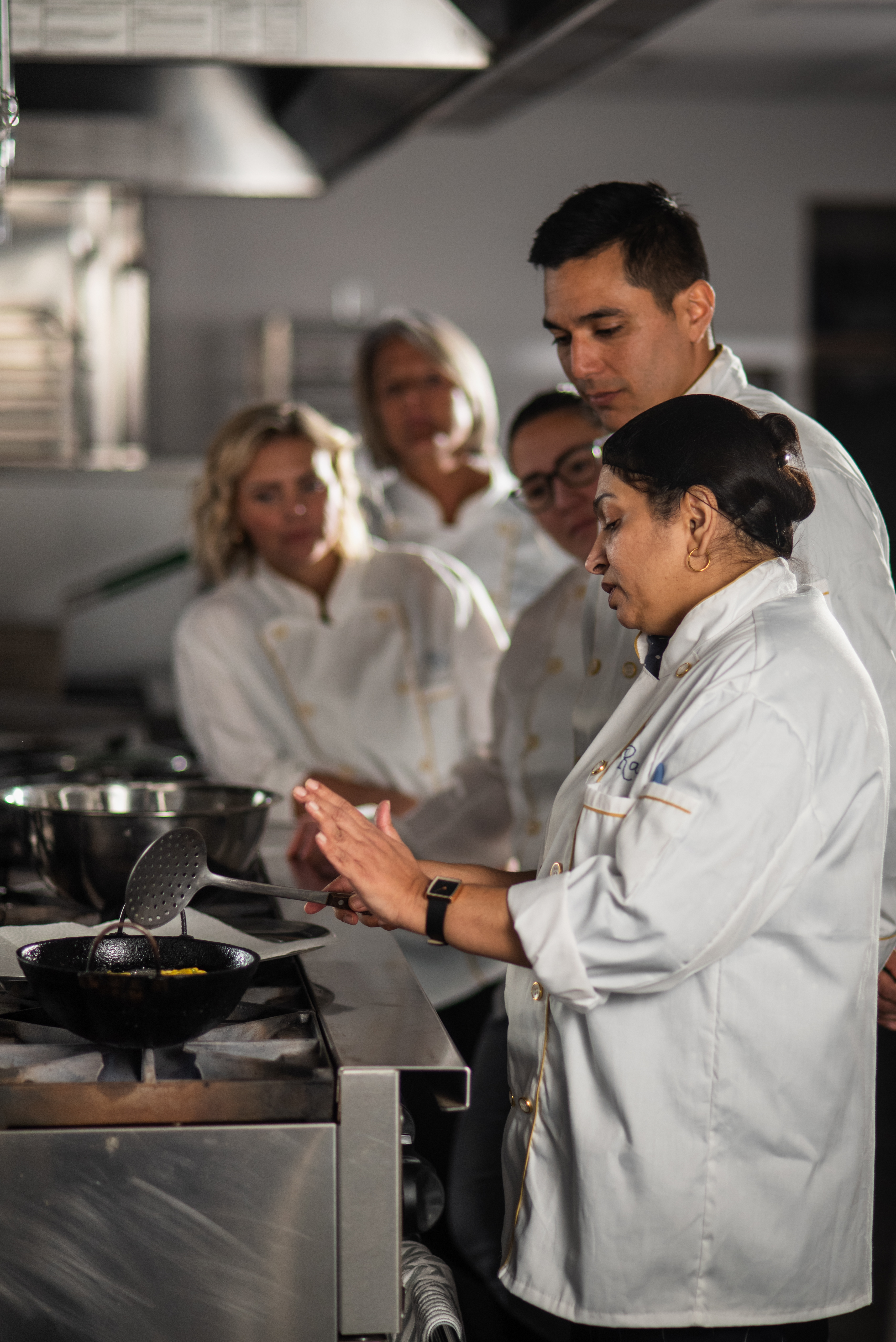 A women in a white jacket instructs people how to cook in front of a stove