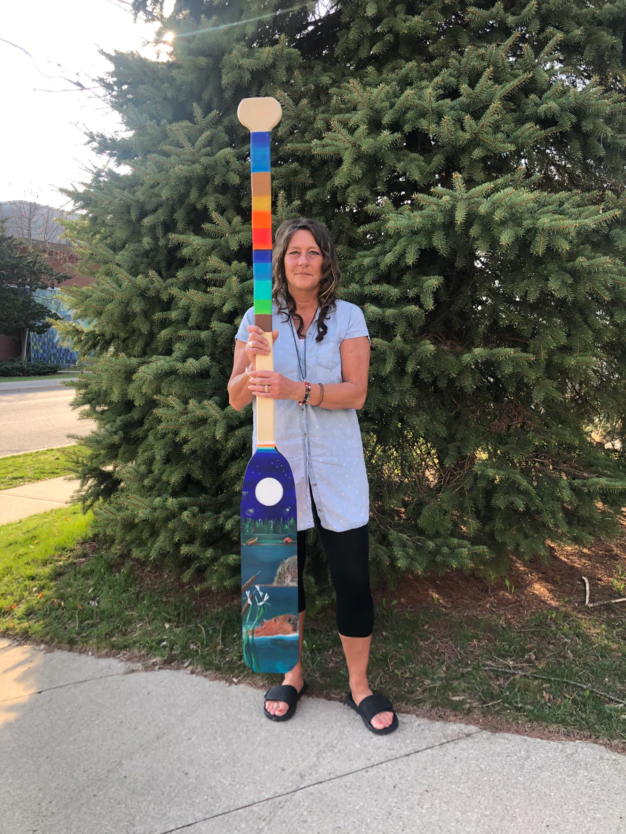 A woman stands in front of a large tree holding a painted paddle