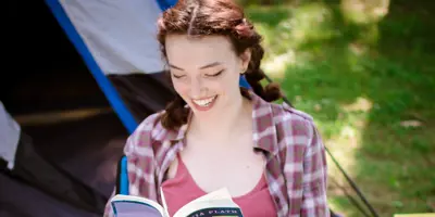 A woman sits in a lawn chair reading a book in front of a blue tent